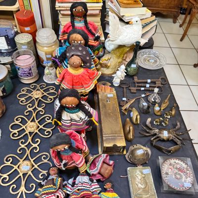 Assortment of vintage collectibles including handmade Mexican rag dolls, brass figurines, old keys, candles, and stacks of books.