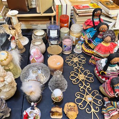 Assorted tabletop decor including Mexican rag dolls, jar candles, angel figurines, and a gold-tone metal wall hanging.