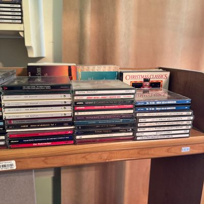 A collection of music CDs in jewel cases stacked on a wooden shelf, featuring artists like The Beatles, Kenny G, Yanni, and various...