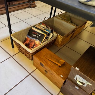 Vintage wooden shoe shine box with footrest alongside woven storage baskets containing books and papers, displayed on a tiled floor.