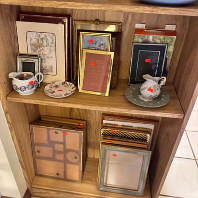 Assorted vintage picture frames in brass, wood, and metal finishes, displayed alongside decorative porcelain plates and small pitchers on...