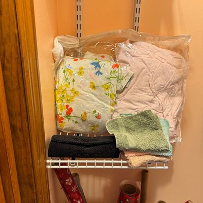 Assorted household linens, including a floral sheet set and pink blanket in plastic storage bags, stacked with washcloths and a black...