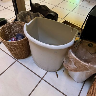Assorted household bins, including a woven wicker basket containing small hand weights and two plastic wastebaskets, sitting on a tiled...