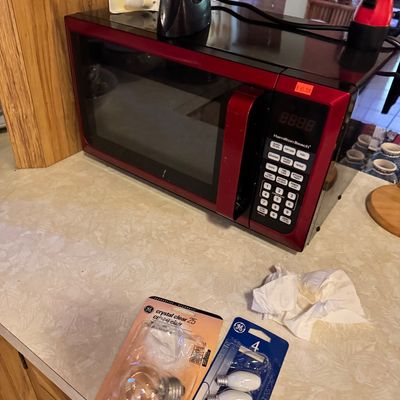 Hamilton Beach countertop microwave in metallic red and black finish, shown alongside packaged lightbulbs on a kitchen counter.