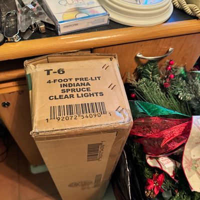 Boxed 4-foot pre-lit Indiana Spruce artificial Christmas tree with clear lights, shown alongside festive holiday garlands and ribbons.