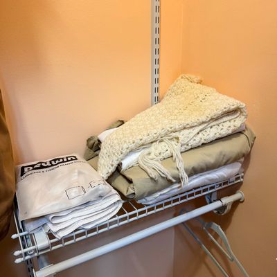 Assorted linens on a wire closet shelf, including a cream-colored crocheted throw blanket with fringe, folded sheets, and a packaged...
