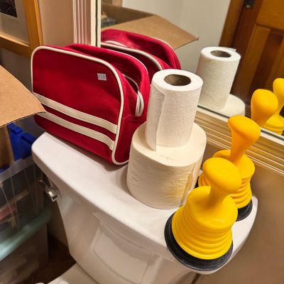 Assorted bathroom items including a red zippered travel bag with white stripes, two rolls of toilet paper, and two yellow mini plungers.