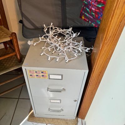 Two-drawer metal filing cabinet in light gray, topped with a tangled strand of white Christmas lights.