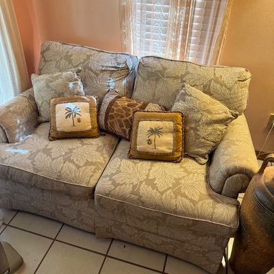 Upholstered loveseat featuring a beige and white leaf pattern, styled with matching throw pillows and accent pillows with palm tree and...