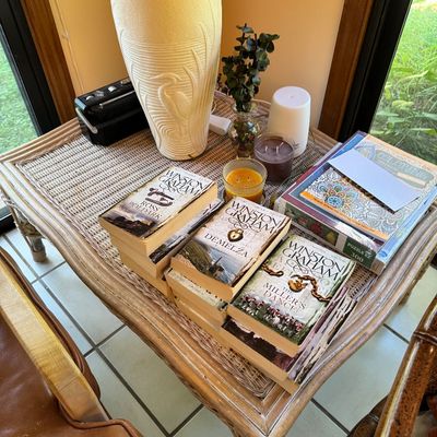 Wicker and rattan side table holding a collection of Winston Graham Poldark novels, a textured ceramic table lamp with a heron design,...