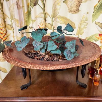 Decorative shallow bowl with a woven rattan rim and leather-like interior, supported by a black wrought iron stand and filled with faux...