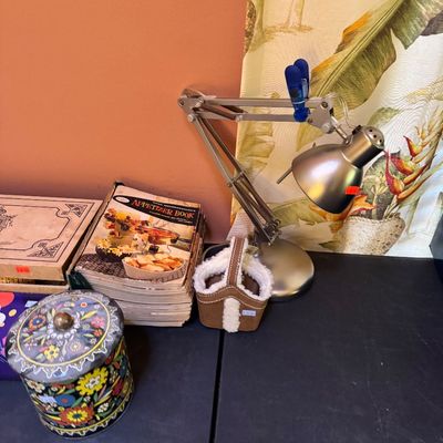 Vintage metal architect desk lamp alongside a stack of cookbooks, including a Good Housekeeping Appetizer Book, and a colorful floral tin...