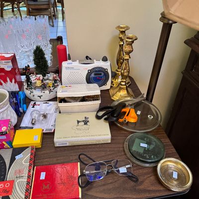 Assorted household items on a table, including brass candlesticks, an Emerson weather radio, sewing thread, scissors, and eyeglasses.