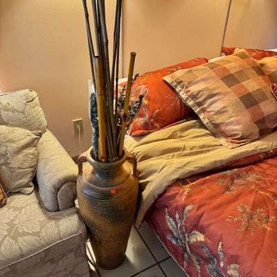 Large rustic ceramic floor vase with handles, filled with decorative bamboo stalks, shown alongside a bed and armchair.