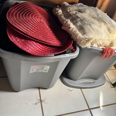 Two grey Sterilite plastic storage bins filled with household items, including red woven placemats and fringed textiles.