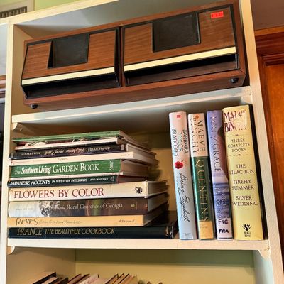 Vintage woodgrain 8-track tape storage unit sitting on a bookshelf filled with various fiction and non-fiction books.