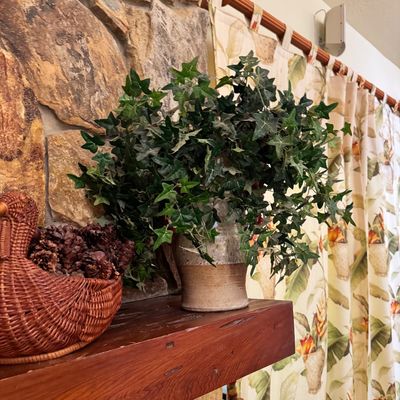 Faux ivy plant in a rustic ceramic pot, displayed on a wooden mantel alongside a woven wicker basket filled with pinecones.