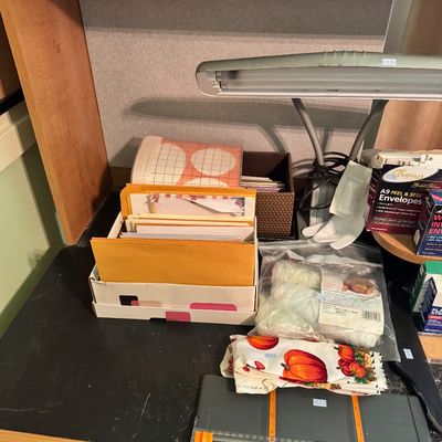 Assorted office and crafting supplies, including envelopes, a paper trimmer, a desk lamp, and a bag of yarn, arranged on a black desk...