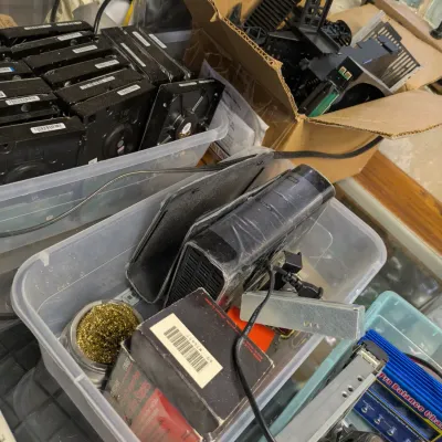 Sale Photo Thumbnail #113: Assorted electronics and computer parts, including a stack of hard drives in a plastic bin, a cardboard box of plastic components, and various cables and accessories.
