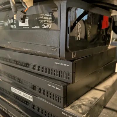 Sale Photo Thumbnail #107: Stack of dusty networking equipment including an APC Back-UPS Pro 1000S uninterruptible power supply resting on top of three Cisco 4300 Series routers.