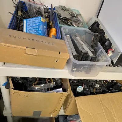 Sale Photo Thumbnail #115: Assorted computer parts, cables, and electronic accessories organized in cardboard boxes and plastic bins on a white shelving unit.
