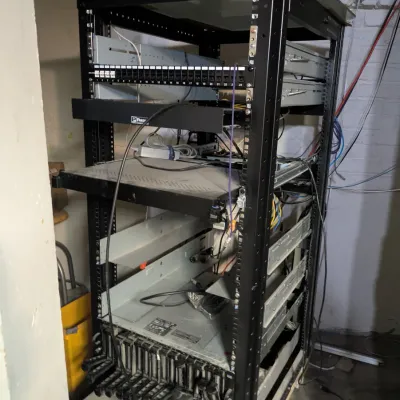 Sale Photo Thumbnail #114: Black metal server rack containing various networking equipment, patch panels, and server chassis, showing significant dust and wear.