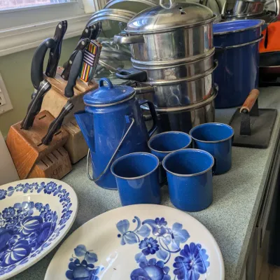 Sale Photo Thumbnail #125: Assorted kitchenware including blue enamelware mugs and coffee pot, stainless steel steamer pots, ceramic plates with blue floral patterns, and a wooden knife block set.
