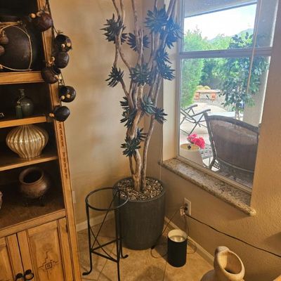 Tall artificial succulent tree in a grey cylindrical planter, accompanied by a black metal plant stand and a rustic ceramic jug.