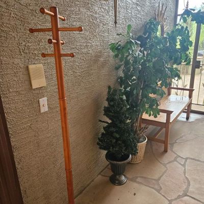 Vintage wooden coat rack with a round base and multiple pegs, shown alongside potted artificial plants and a wooden bench 