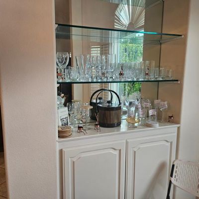 Built-in white bar cabinet with mirrored back and glass shelves, displaying an assortment of glassware, wine glasses, and a mid-century...