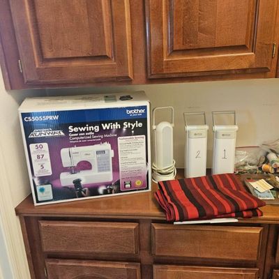 Brother CS5055PRW Project Runway computerized sewing machine in its original box, displayed alongside sewing accessories, fabric, and...