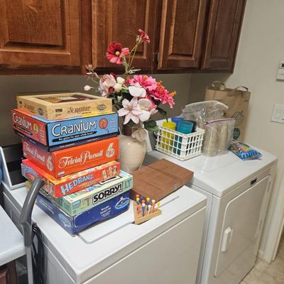 A stack of classic board games including Scrabble, Cranium, Trivial Pursuit, Hedbanz, and Sorry!, displayed alongside household items in...
