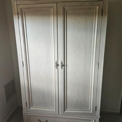 Traditional wooden armoire with a whitewashed finish, featuring two paneled doors, a lower drawer with brass pulls, and a classic crown...