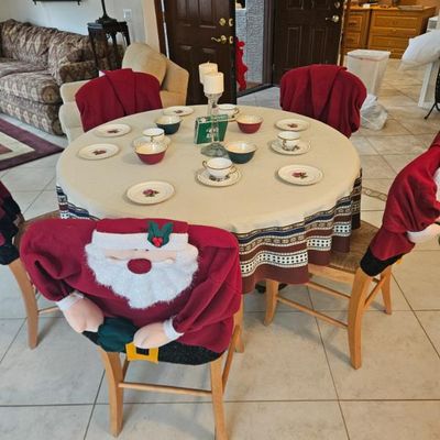 Round wooden dining table set with four chairs, featuring festive Santa Claus chair back covers and a holiday-themed table setting.