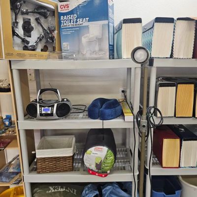 Assorted household items on plastic shelving, including a CVS raised toilet seat in its box, a portable CD player/radio, a blue neck...