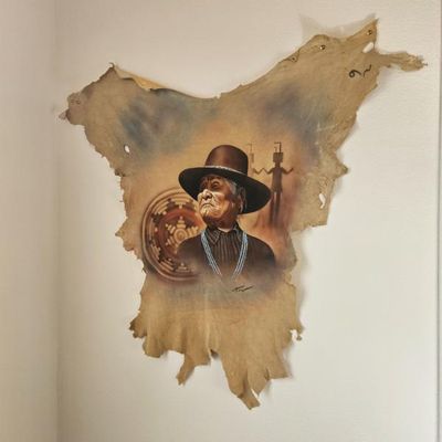 Native American portrait painted on a shaped leather hide, featuring an elder in a hat with traditional motifs in the background.