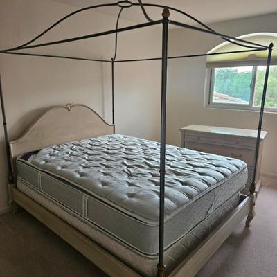 French Provincial style four-poster canopy bed with a whitewashed wooden frame, metal canopy rails, and matching three-drawer dresser in...