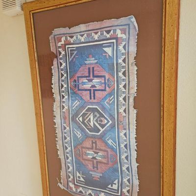 Framed Southwestern-style textile art featuring geometric patterns in blue, pink, and white, mounted on a brown mat inside a gilded...