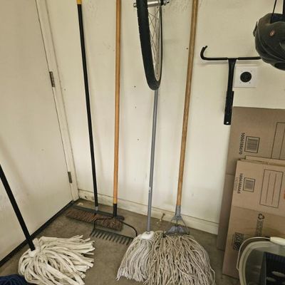 Assorted cleaning and garden tools, including two string mops, a push broom, and a metal rake, alongside flat-packed Home Depot moving...