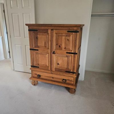 Rustic pine armoire featuring paneled double doors with black iron strap hinges and a single bottom drawer with ring pulls.