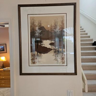 Framed landscape print depicting a serene lake and mountain scene in muted brown and cream tones, housed in a dark wood frame with a wide...