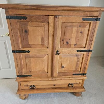 Rustic pine armoire featuring black iron strap hinges, a lower drawer with ring pulls, and bun feet.