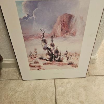 Framed desert landscape print featuring Joshua trees and a distant mesa under a stormy sky, presented in a simple metal frame with a wide...