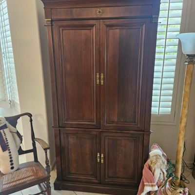 Traditional dark wood corner armoire featuring brass hardware and a pediment top, flanked by a cane-seat rocking chair and holiday...
