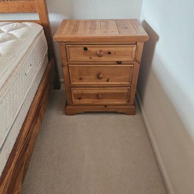 Rustic knotty pine three-drawer nightstand with wooden knobs, shown next to a matching wooden bed frame.
