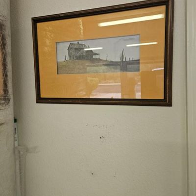 Framed landscape print depicting a rustic house on a hill, matted in yellow and housed in a dark wood frame.