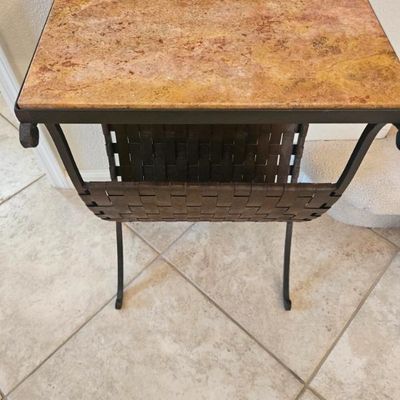 Wrought iron side table featuring a polished travertine stone top and an integrated woven leather magazine sling underneath.