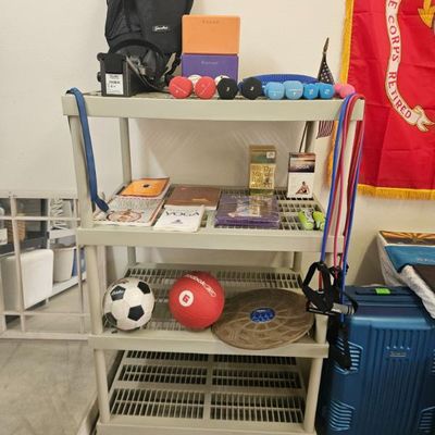 Four-tier plastic utility shelf holding various fitness equipment, including hand weights, yoga blocks, exercise books, a soccer ball,...