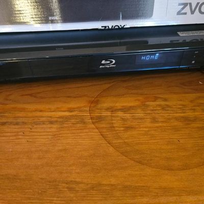 Sony Blu-ray Disc player shown powered on and resting on a wooden surface, with a ZVOX box visible in the background.