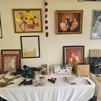 Assorted household items, framed family photos, children's artwork, inline skates, and tools displayed on tables for sale.